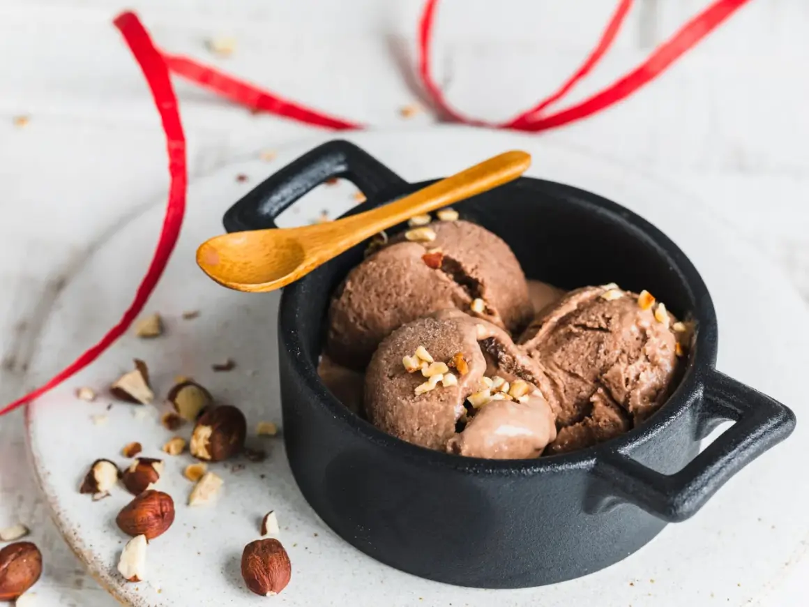 Schokoladiges Nutella-Eis mit 3 Zutaten, garniert mit gehackten Haselnüssen in einer schwarzen Schale, daneben eine Holz-Löffel und rote Bänder.