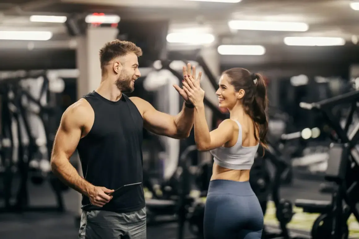 Mann und Frau geben sich im Fitnessstudio einen High Five. Er hält ein Tablet, vielleicht für seine persönliche Coach Ausbildung.