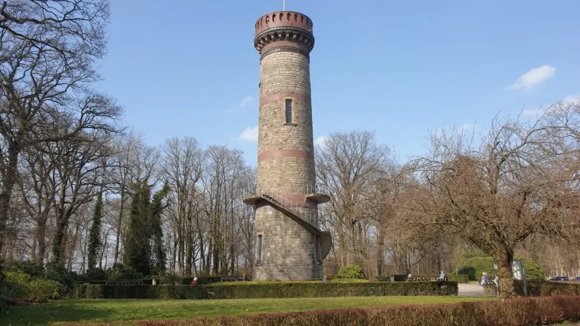 Der Toelleturm in Wuppertal, ein steinerner Aussichtsturm, ragt vor kahlen Bäumen und blauem Himmel auf. Eine Wendeltreppe führt zur Aussichtsplattform.