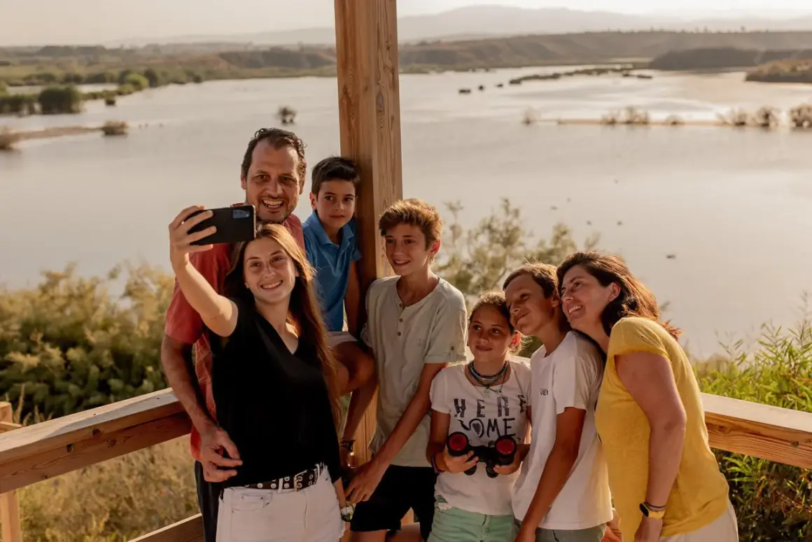 Familia sonriente se toma un selfie con un lago de fondo. Un día perfecto en la playa de Vera para niños, explorando la naturaleza.