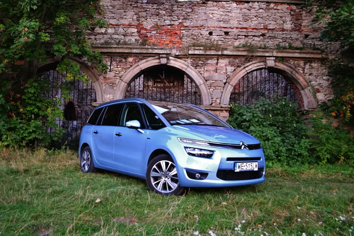 Błękitny Citroen C4 Grand Picasso 2.0 HDI zaparkowany na trawie przed zabytkowym murem z cegły i kamienia.
