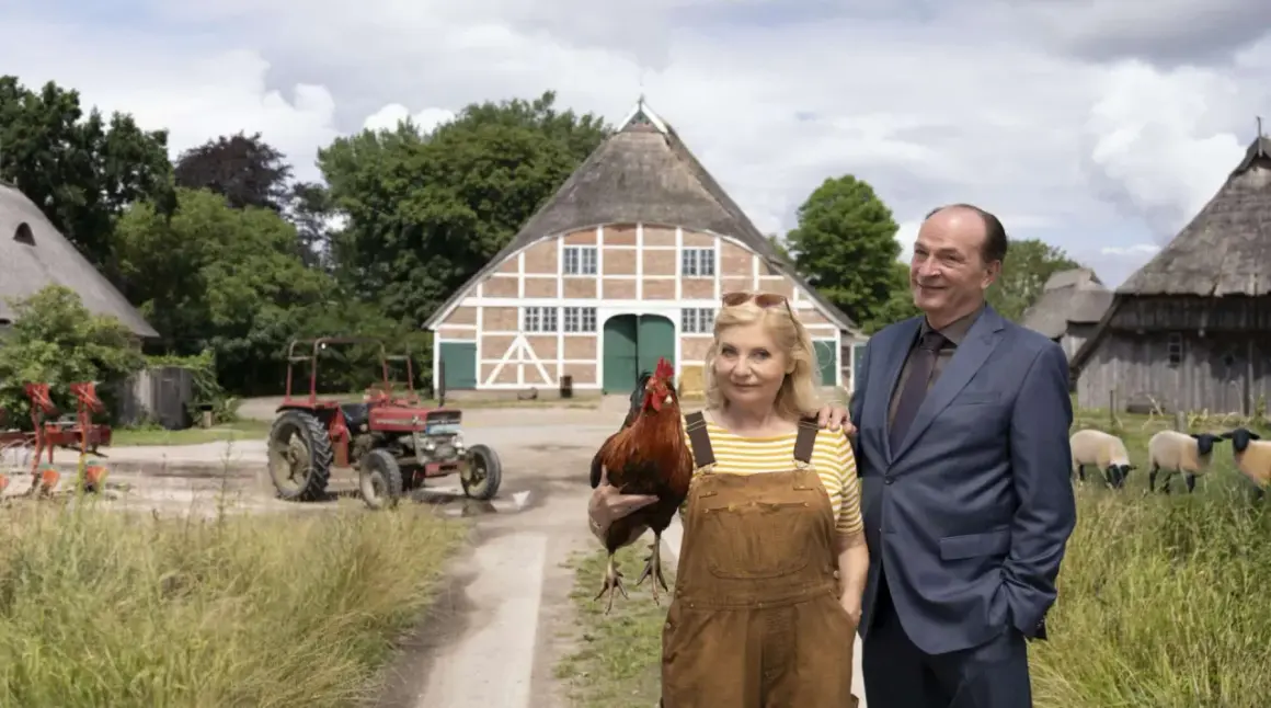 Ein Mann im Anzug und eine Frau mit Latzhose und Huhn stehen vor einem Bauernhaus. Im **weiten Land** grasen Schafe, ein Traktor steht bereit.