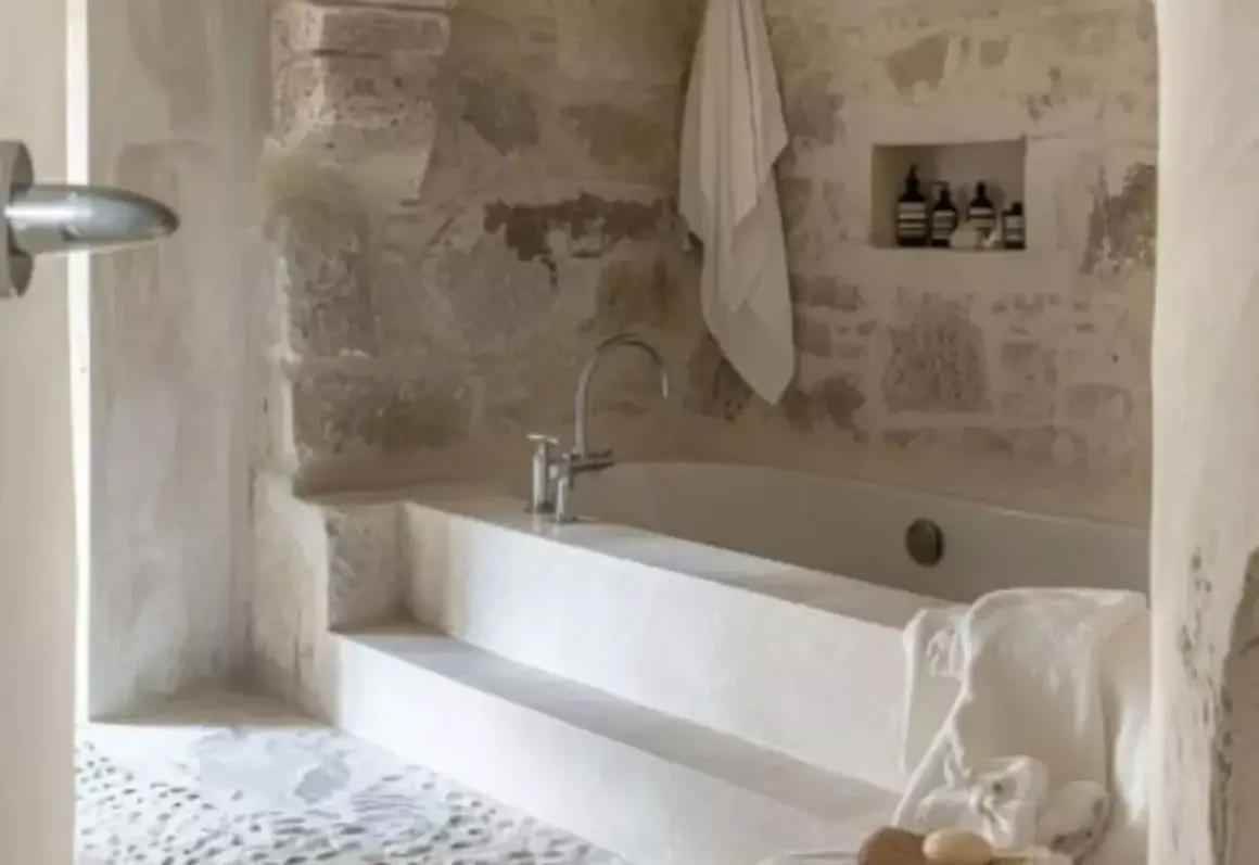 Une **salle de bain méditerranéenne** avec baignoire en pierre brute, robinetterie moderne et serviette blanche suspendue.