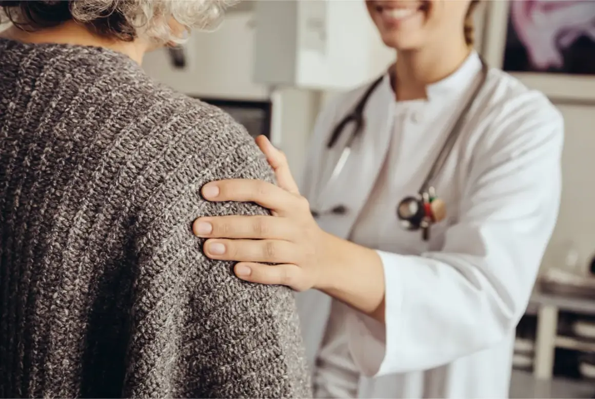 Une doctoresse réconforte une patiente âgée, lui posant une main sur l'épaule. Elle rassure la patiente qui a peur de se faire pipi dessus.