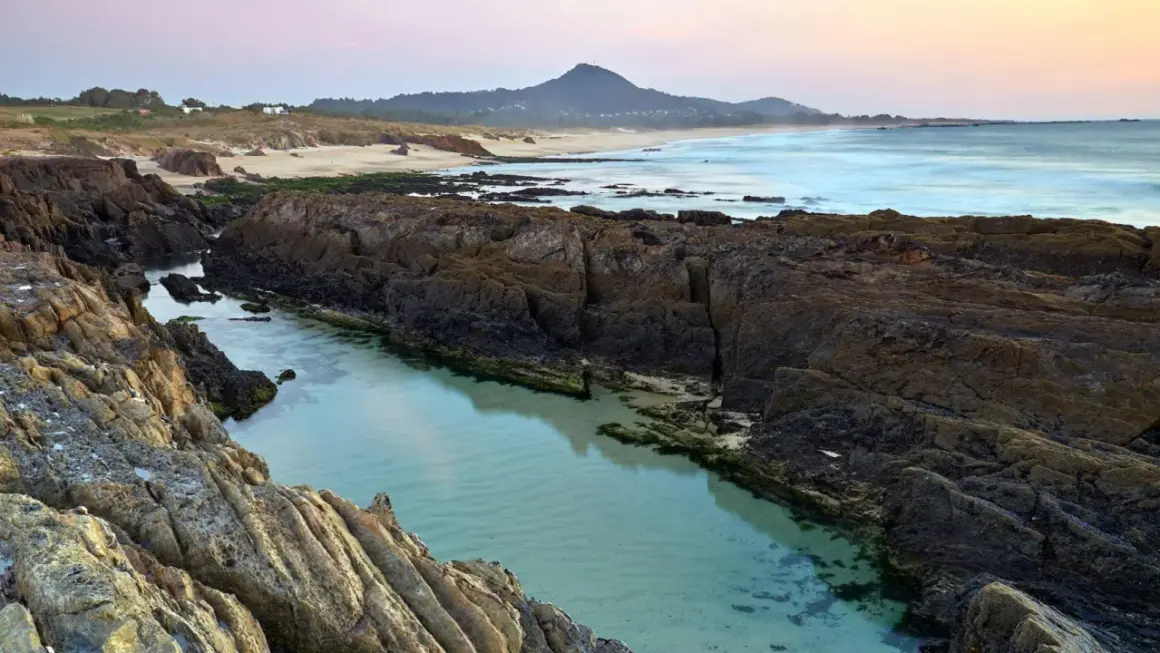 Aguas cristalinas en la **playa das Furnas**, entre rocas volcánicas y arena dorada, con el mar al fondo y una montaña.
