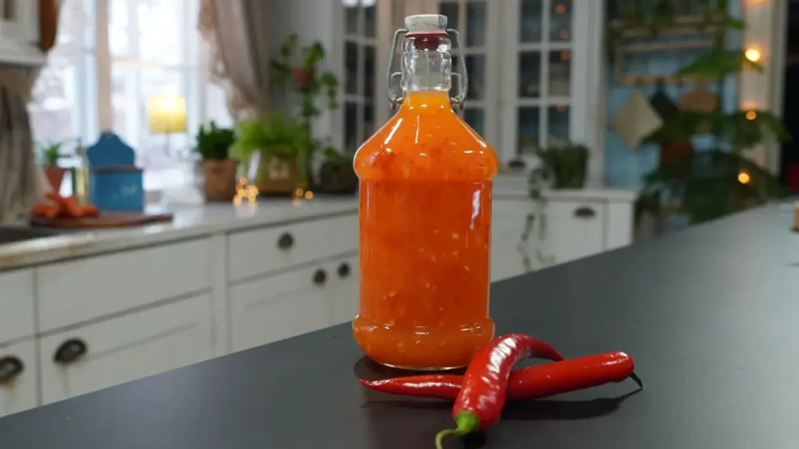 En flaska med hemmagjord sweet chili sås står på en mörk bänk, med två röda chilifrukter framför.