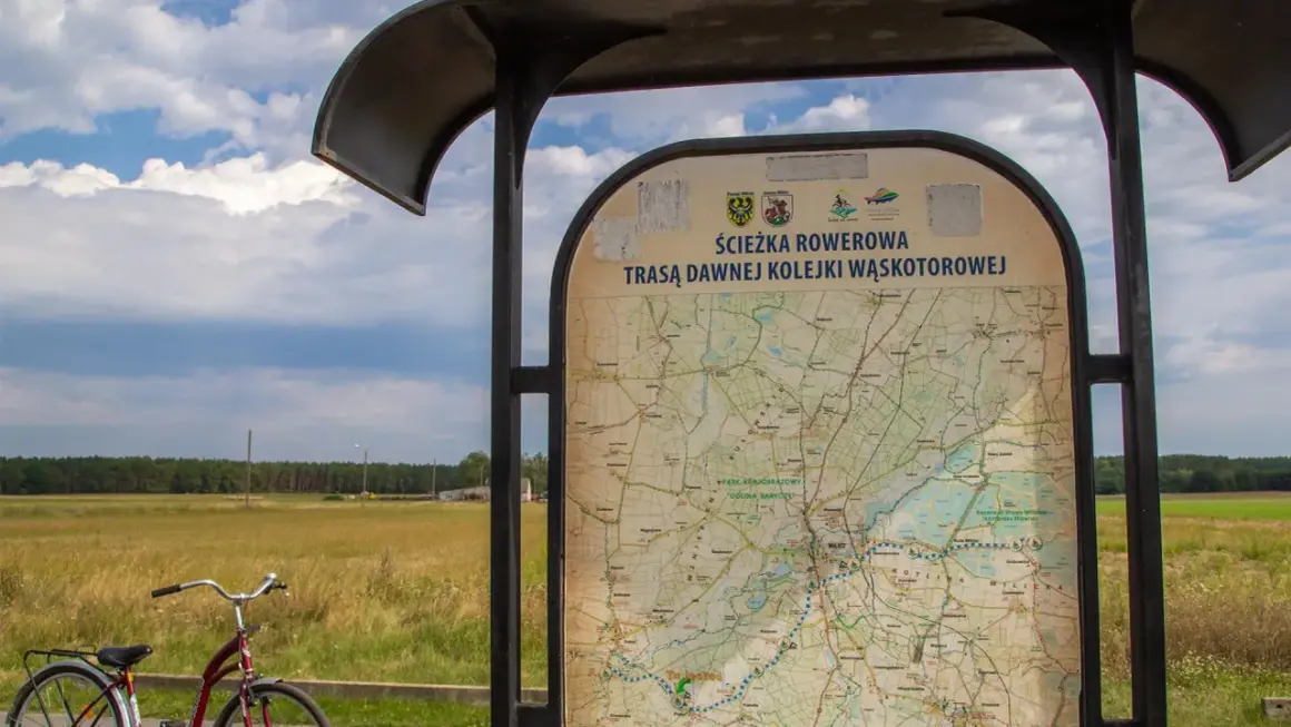Mapa informuje o ścieżce rowerowej trasą dawnej kolejki wąskotorowej. Obok stoi rower.