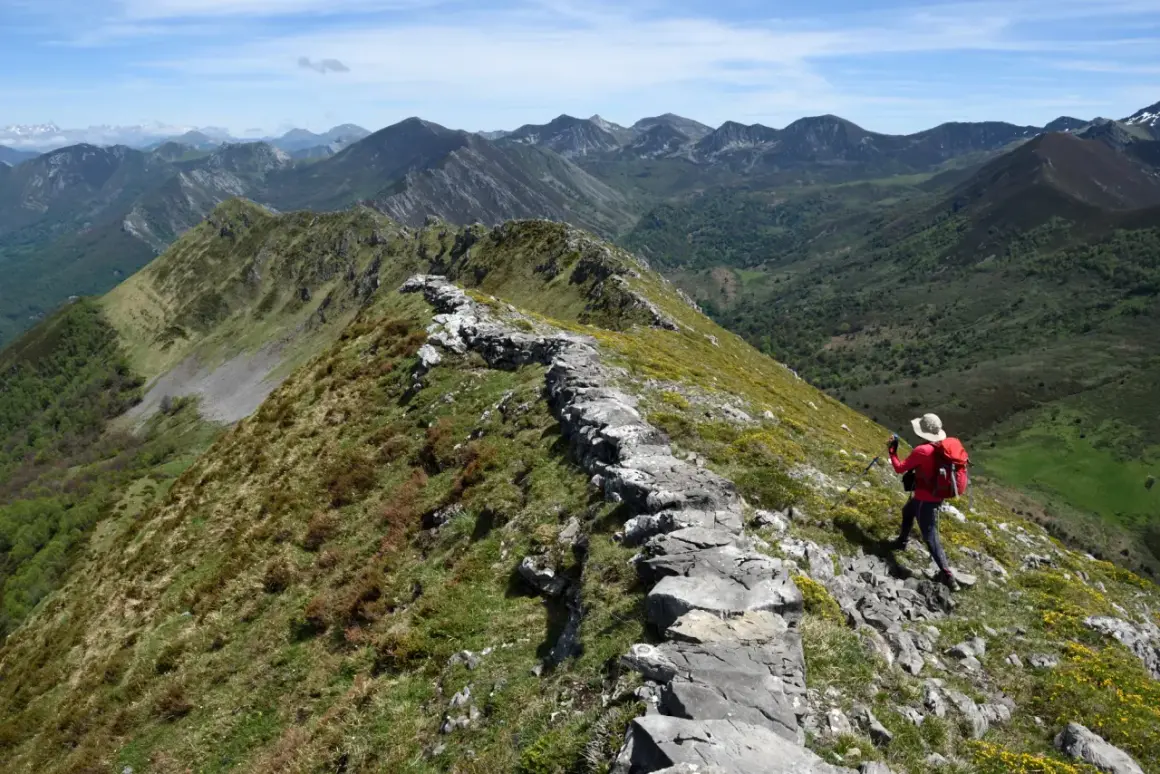 Senderista en cresta rocosa, disfrutando de vistas panorámicas de montañas verdes. Un ejemplo de las mejores rutas de senderismo en España.