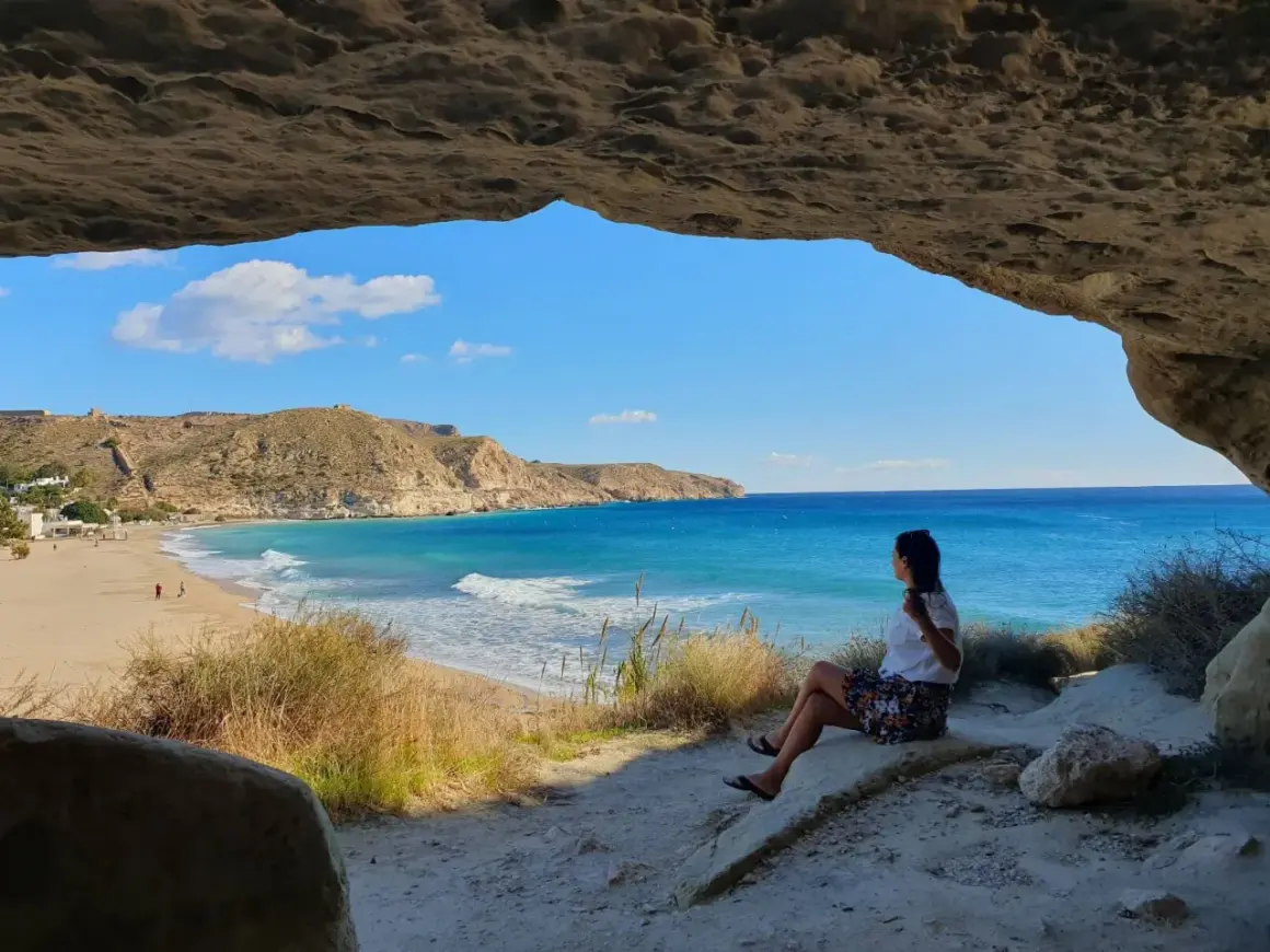 Mujer sentada en una cueva, contemplando la playa y el mar azul. Un lugar idílico que ver cerca de Cabo de Gata.