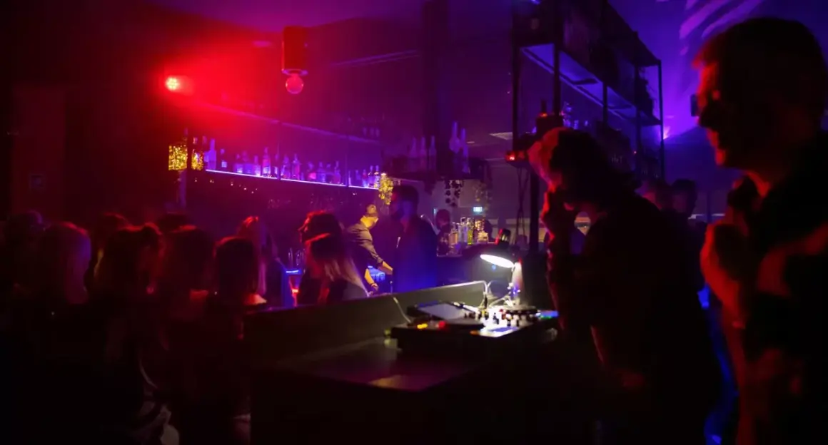 Un hot night club con luci rosse e viola, gente che balla e un DJ al lavoro. L'atmosfera è vibrante e piena di energia.