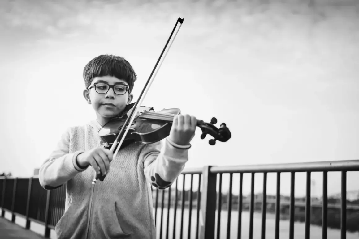 Ein Junge mit Brille spielt konzentriert Geige. Er steht an einer Brüstung, die Musik erklingt im Freien.