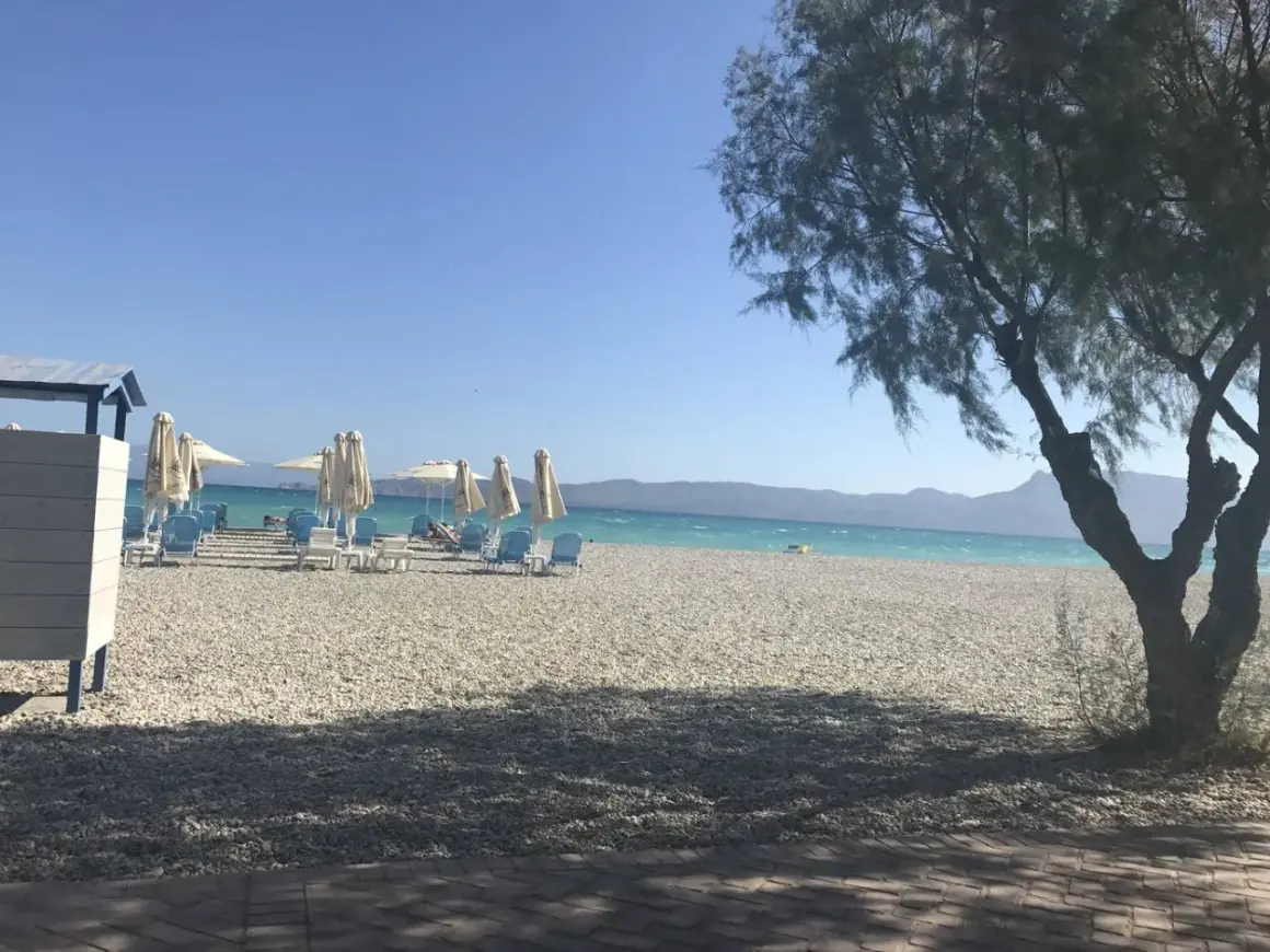 Słoneczny dzień na plaży Alkyon. Białe leżaki i parasole czekają na plażowiczów. W tle turkusowe morze i góry.