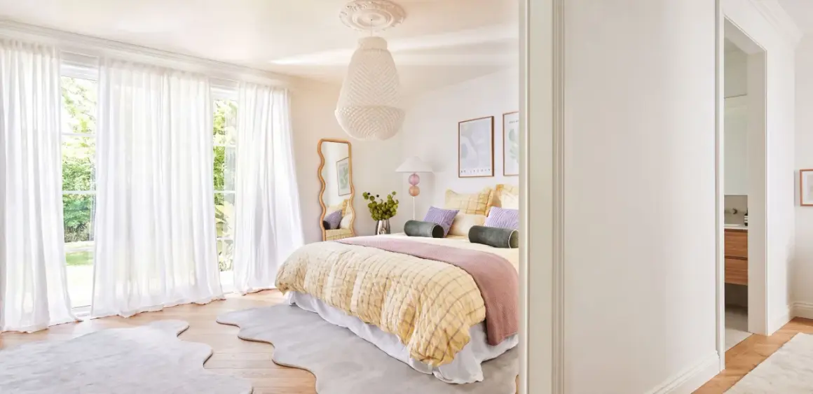 Ein geschwungener Spiegel im Schlafzimmer reflektiert das helle Zimmer mit Bett, weißen Vorhängen und Blick ins Grüne.