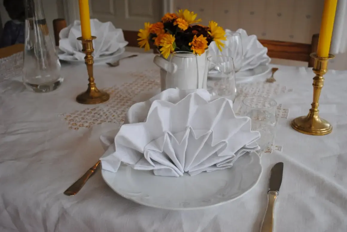 Vackert dukat bord med vita, vikta servetter i glas. Guldljusstakar och blommor skapar en festlig stämning.