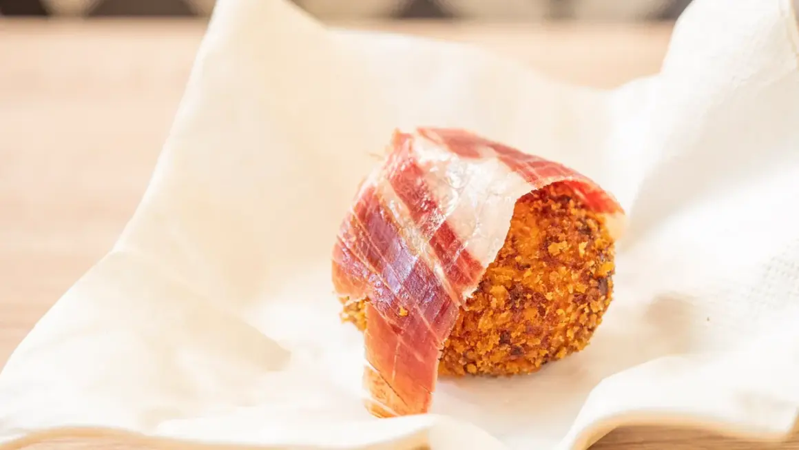 Croquetas de jamón crujientes, envueltas en una loncha de jamón serrano, servidas sobre papel blanco.