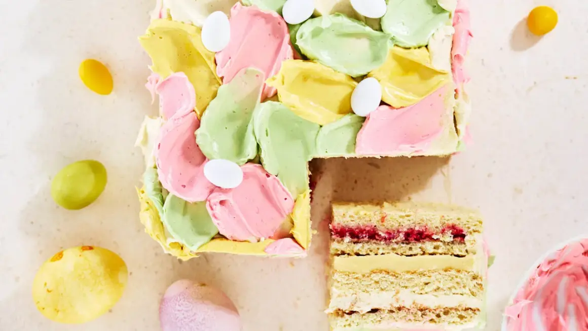En vacker vit choklad tårta med pastellfärgad frosting och påskägg. En bit är uppskuren och visar lager av kräm och sylt.