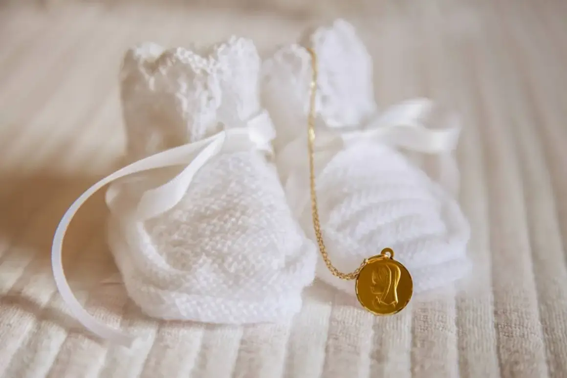 Douces chaussons blancs tricotés, ornés de rubans, avec une médaille dorée de la Vierge. Un symbole de marraine sans baptême, plein d'amour.