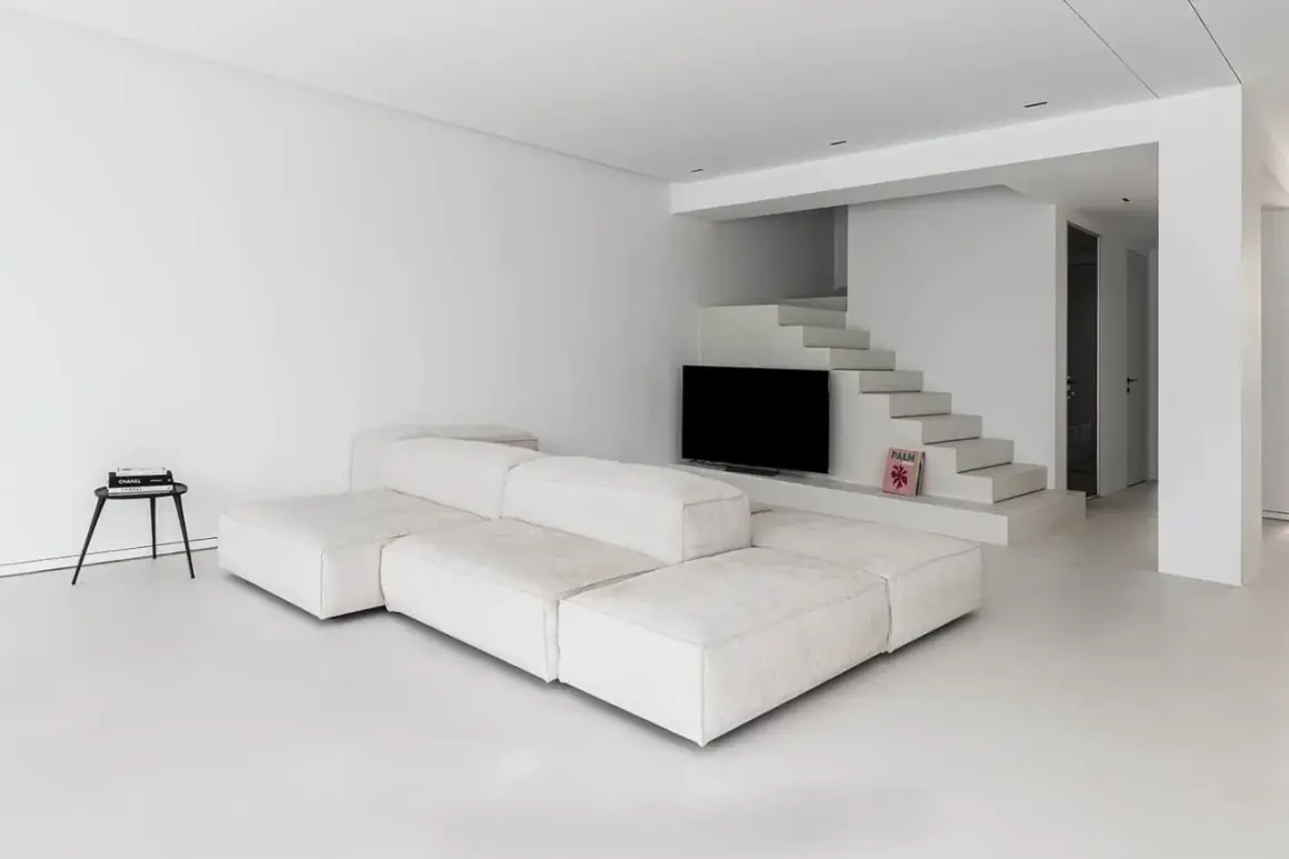 Soggiorno minimalista con divano modulare bianco e pavimento in resina epossidica opaca. Scale moderne conducono a un'area con TV.