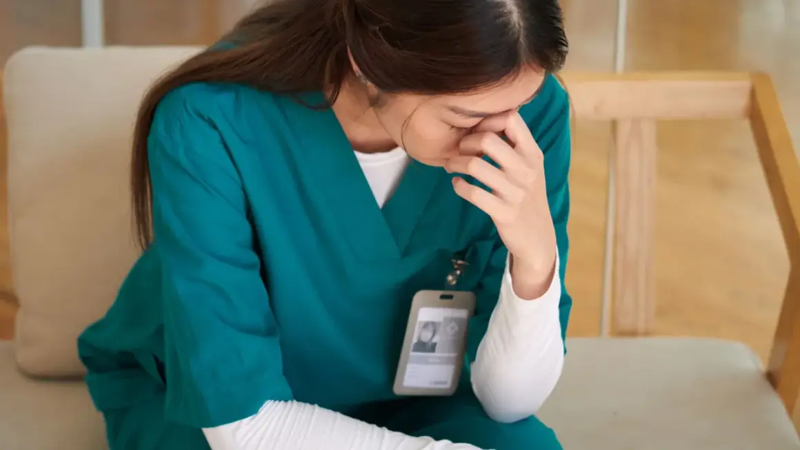 Une infirmière en blouse verte, le visage caché dans ses mains, semble lutter pour trouver son équilibre émotionnel.