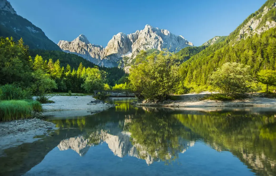 En vacker vy för att vandra i Slovenien. En spegelblank sjö reflekterar berg och skog. En bro leder över vattnet.