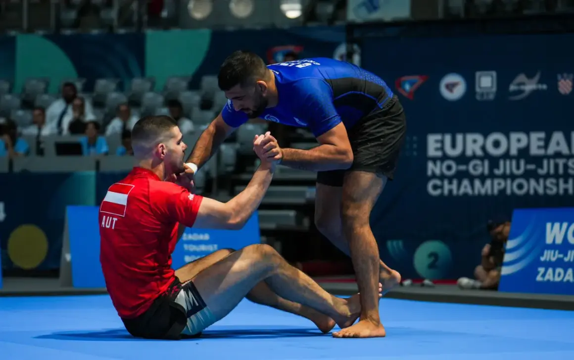 Två män tävlar i ju jitsu. En sitter på mattan i röd dräkt, den andra står i blå dräkt och sträcker ut en hand.