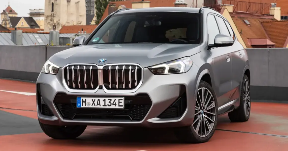 Srebrny x1 bmw z charakterystyczną atrapą chłodnicy i nowoczesnymi światłami, zaparkowany na tle miejskiej panoramy.