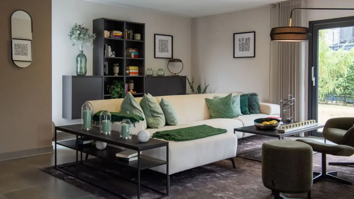 Gemütliches Wohnzimmer mit cremefarbener Couch, grünen Kissen und Decken. Helle Farben dominieren, ergänzt durch dunkle Möbel und eine große Glasfront.