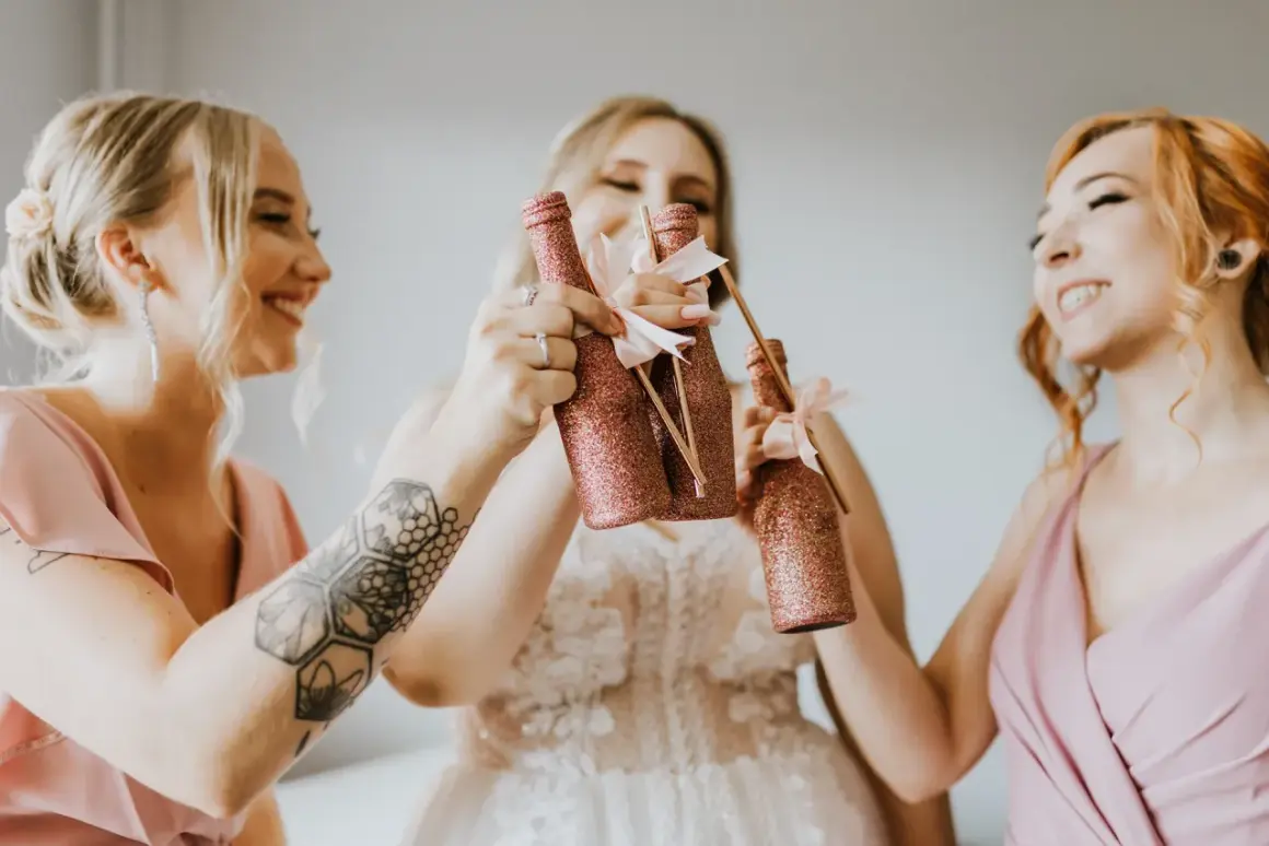 Trzy uśmiechnięte kobiety w różowych sukienkach wznoszą toasty błyszczącymi butelkami. Radosne przygotowania do ślubu.