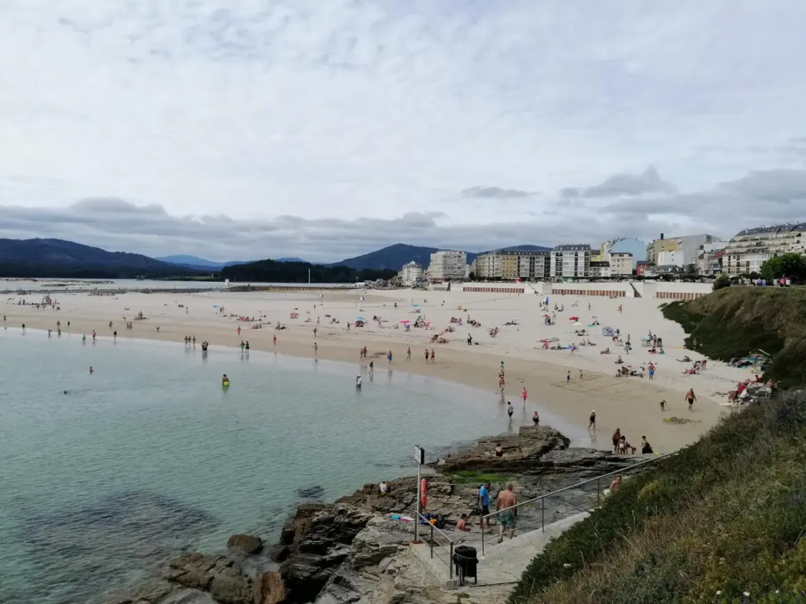 Gente disfruta de un día soleado en la playa de peizas, con aguas cristalinas y edificios al fondo.