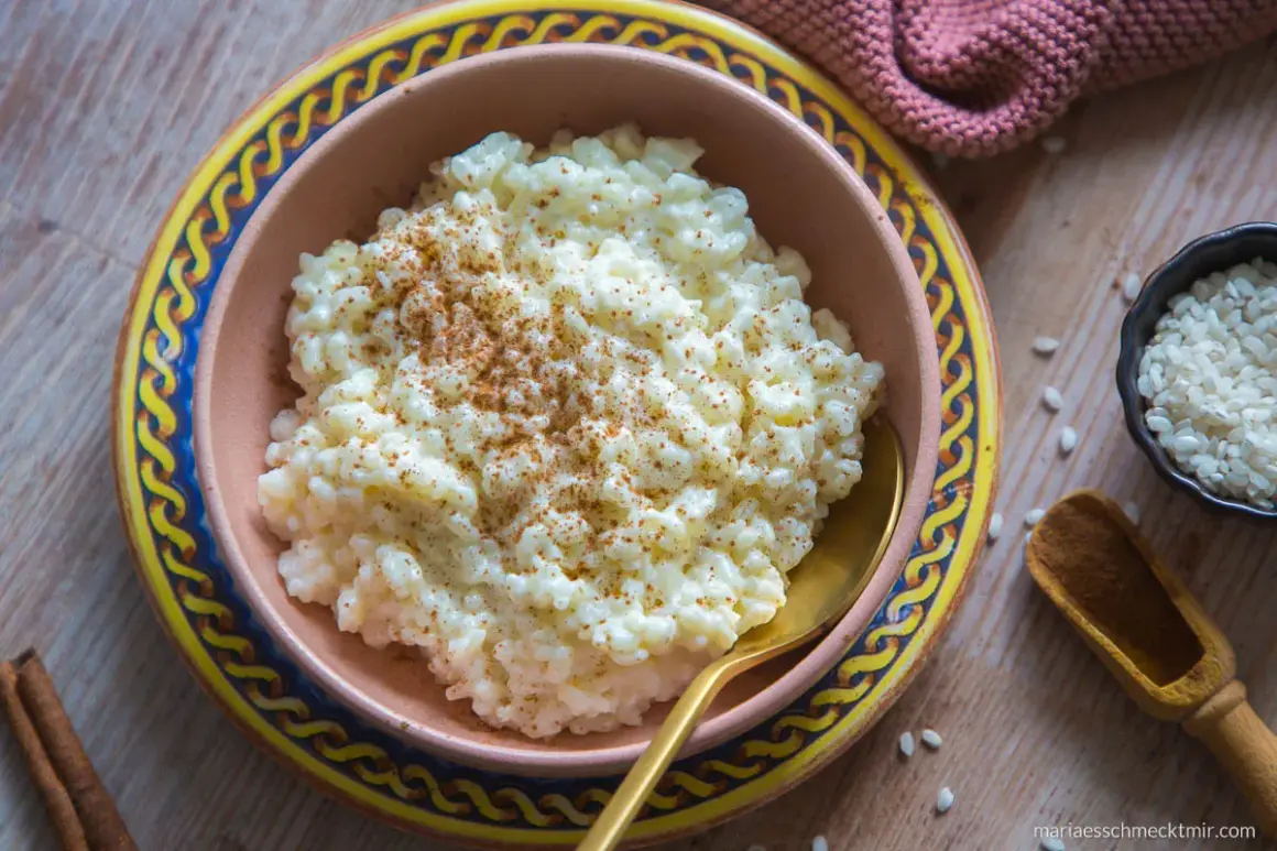Omas Milchreis mit Butter, bestreut mit Zimt, in einer Schüssel auf einem Holztisch. Daneben eine kleine Schale Reis und eine Zimtstange.