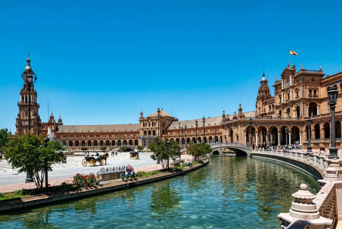 Vacker vy över Plaza de España i Sevilla, Spanien. En hästdragen vagn glider fram längs kanalen under en klarblå himmel.