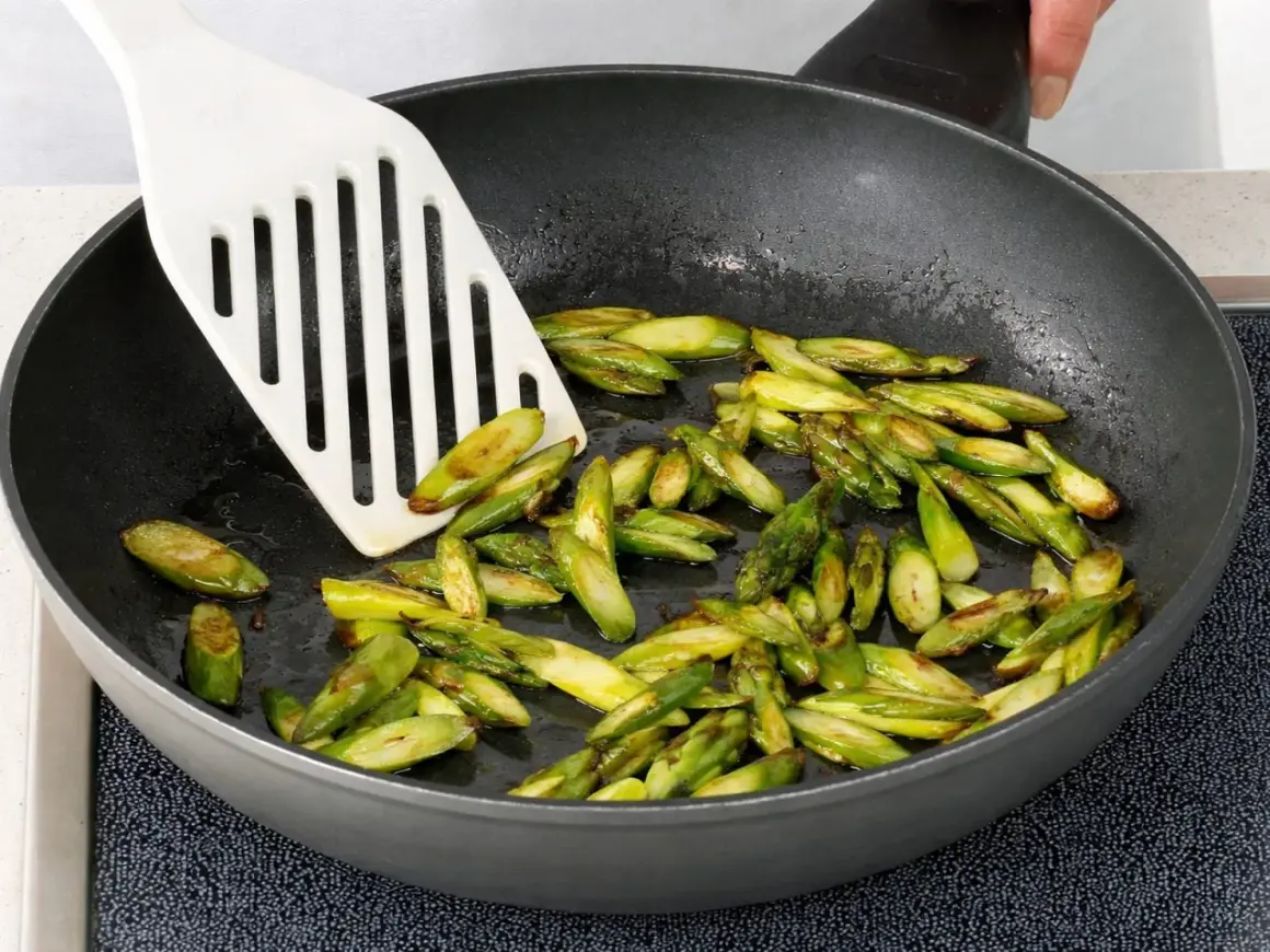 Geschnittener Spargel brät in einer Pfanne. Ein weißer Pfannenwender bewegt die grünen Stücke.