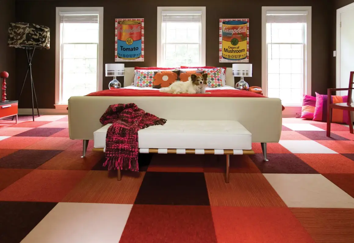 A dog lounges on a bed in a room with a bold, kitsch style. The floor features a checkered pattern of red and brown squares, and the walls are adorned with Campbell's soup can art.