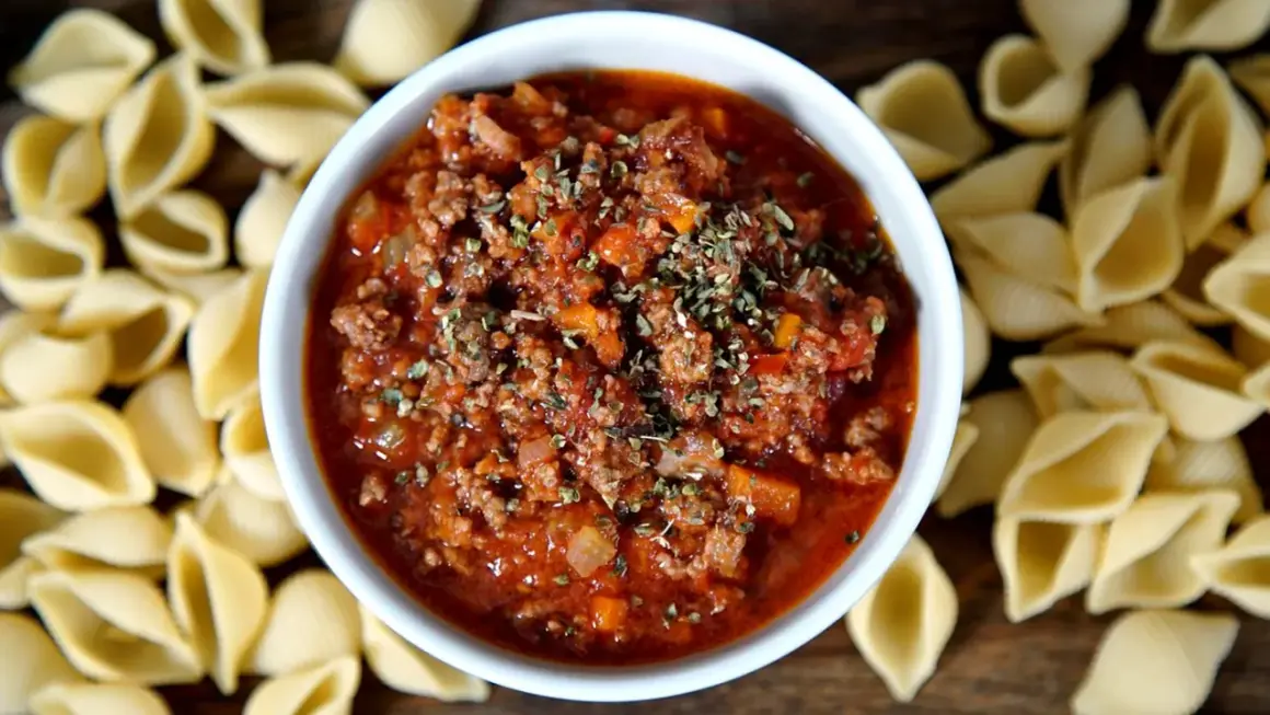 Herzhaftes Bolognese-Rezept mit Hackfleisch und Tomaten, garniert mit Kräutern, umgeben von ungekochten Nudeln.