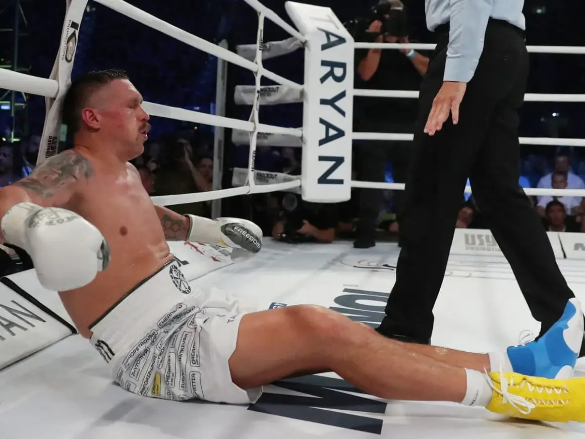 Boxer Oleksandr Usyk sits on the canvas, leaning against the ropes, appearing to recover from a usyk low blow. The referee stands nearby.