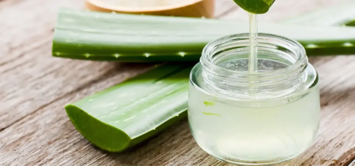 Frisches, reines Aloe Vera Gel tropft aus einem Blatt in ein Glasgefäß. Daneben liegen weitere Aloe Vera Blätter auf Holz.