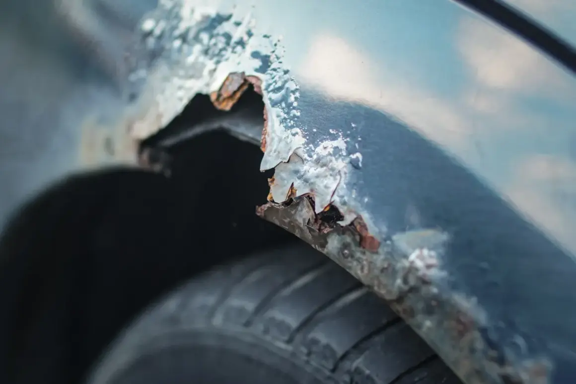 Detaljerad bild av rostig bilskärm. En del av skärmkanten har flagnat och blottar metallen under, redo att laga rost bil. Däcket syns nedtill.