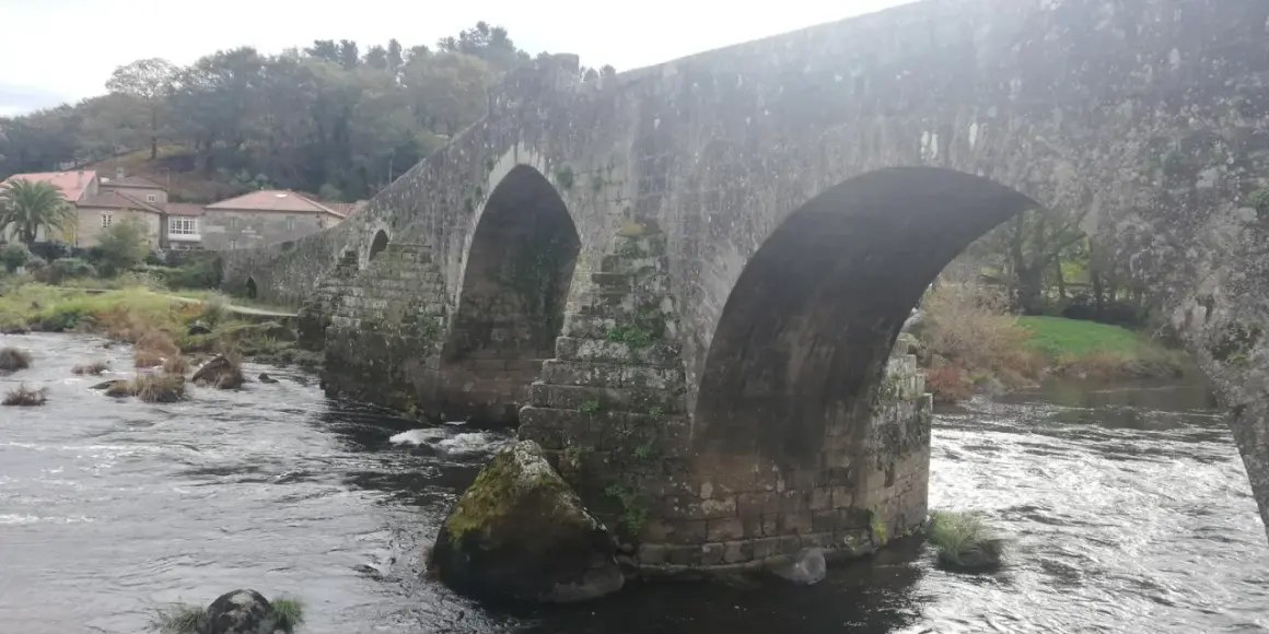 El histórico puente Maceira cruza un río caudaloso, con casas de tejados rojos y árboles frondosos al fondo.