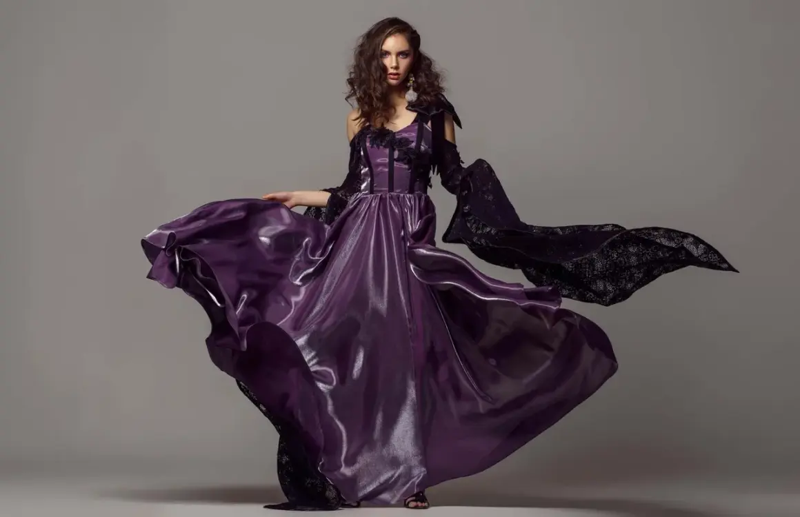 A model in a flowing purple gown and black lace sleeves poses for studio fashion photography.