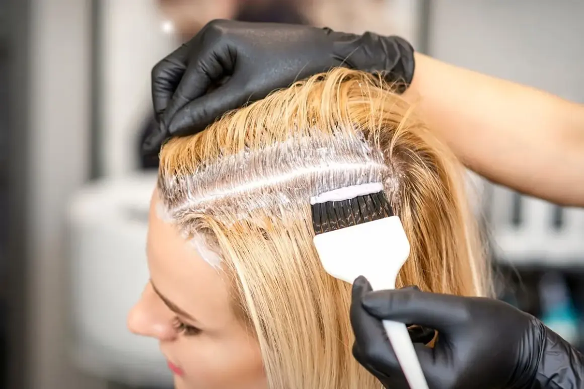 Fryzjerka w czarnych rękawiczkach nakłada farbę na blond włosy, wykonując farbowanie odrostów.