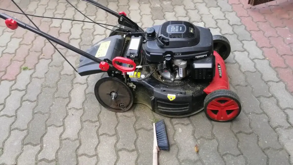 Kosiarka z czerwonymi detalami i solidnymi kółkami do kosiarki, gotowa do pracy na podjeździe z kostki brukowej. Obok leży miotka.