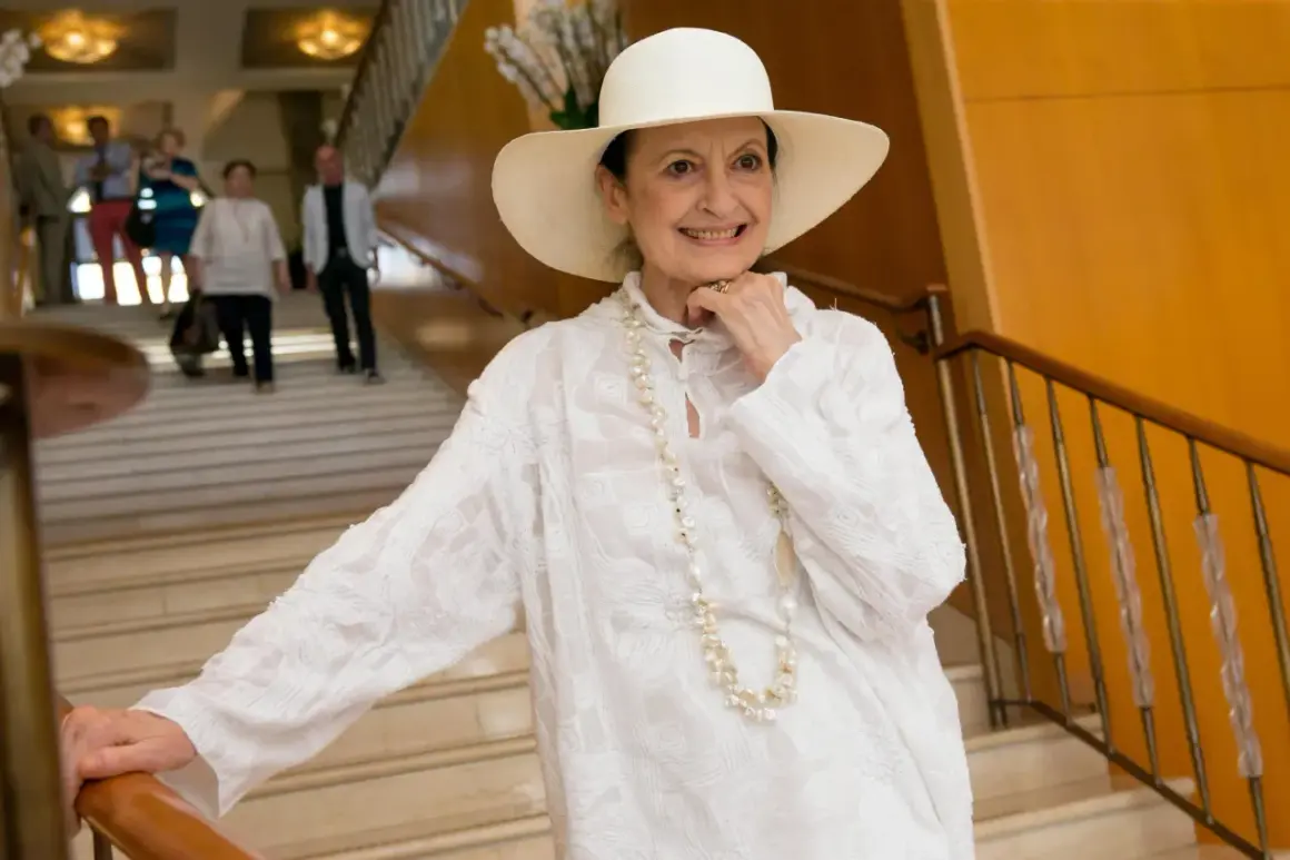 Carla Fracci sorride con un cappello bianco e una collana di perle, elegante su una scala.
