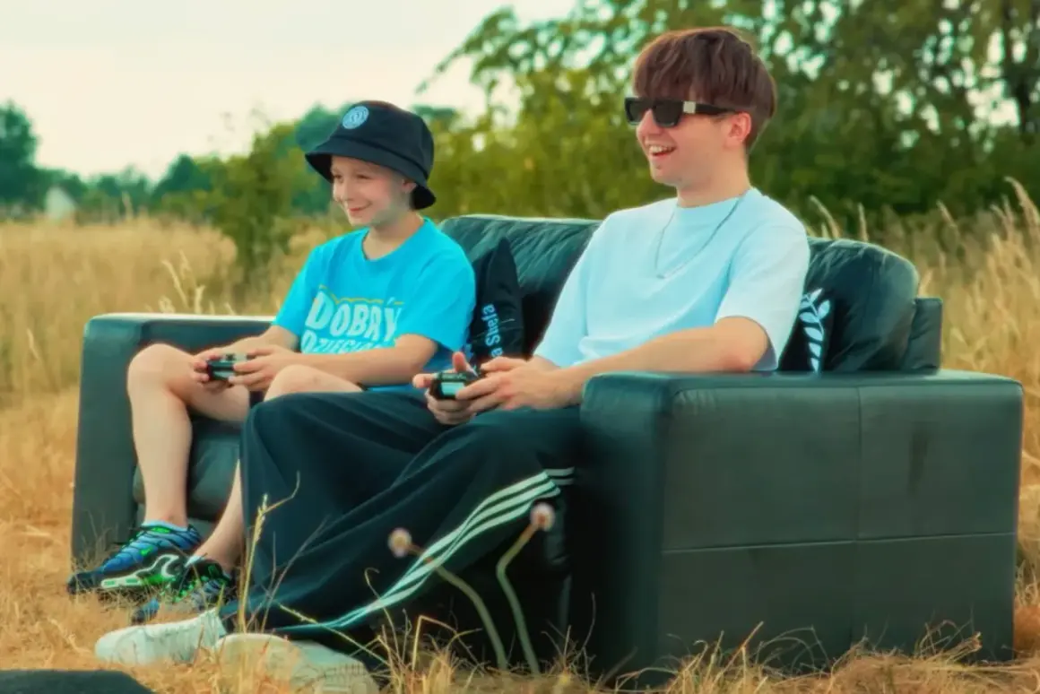 Dwóch chłopców gra w gry wideo na czarnej kanapie na łonie natury. Jeden z nich, młodszy, ma na głowie czarną czapkę typu bucket hat, a drugi, starszy, nosi okulary przeciwsłoneczne. Wyglądają jakby byli na planie teledysku rapera.