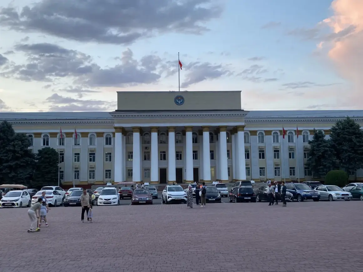 Plac przed budynkiem rządowym w Biszkeku, stolicy Kirgistanu. W tle monumentalna fasada z kolumnami, na dachu flaga. Na placu zaparkowane samochody i spacerujący ludzie.