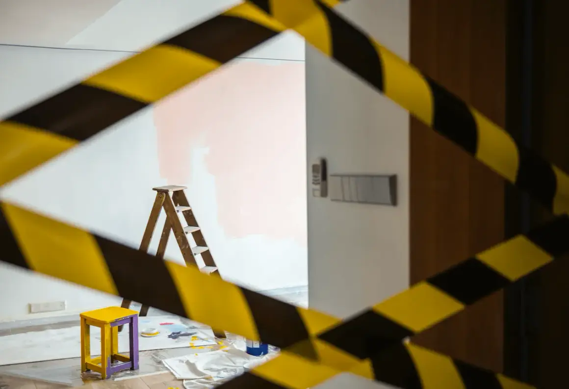Cinta de precaución amarilla y negra rodea una habitación con una escalera, un taburete y pintura rosa en la pared, ilustrando la diferencia entre obras de mejora y obras de conservación.