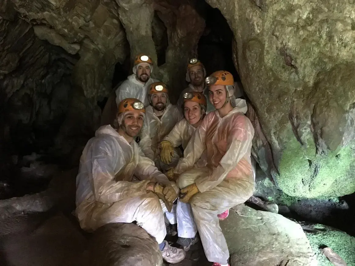 Grupo de exploradores sonríe dentro de la Cova do Rei Centolo, equipados con cascos y trajes protectores.