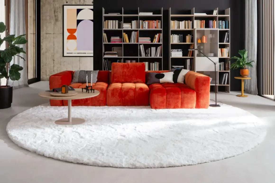 Ein flauschiger weißer Teppich bildet die Basis für das orangefarbene Sofa. So lässt sich der Teppich unter dem Sofa richtig platzieren.