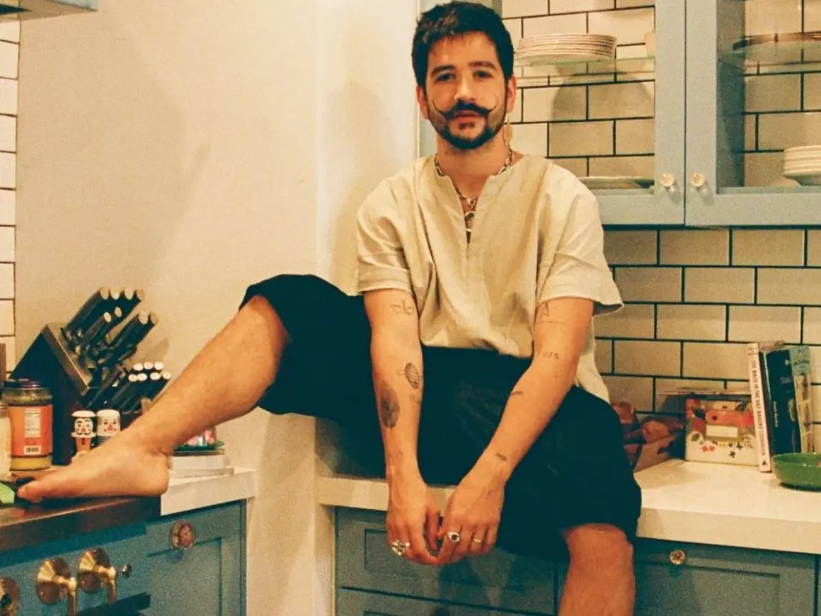 Camilo Alonso Vega, con su característico bigote, posa relajado en una cocina. Sus tatuajes y anillos añaden un toque personal a su estilo.