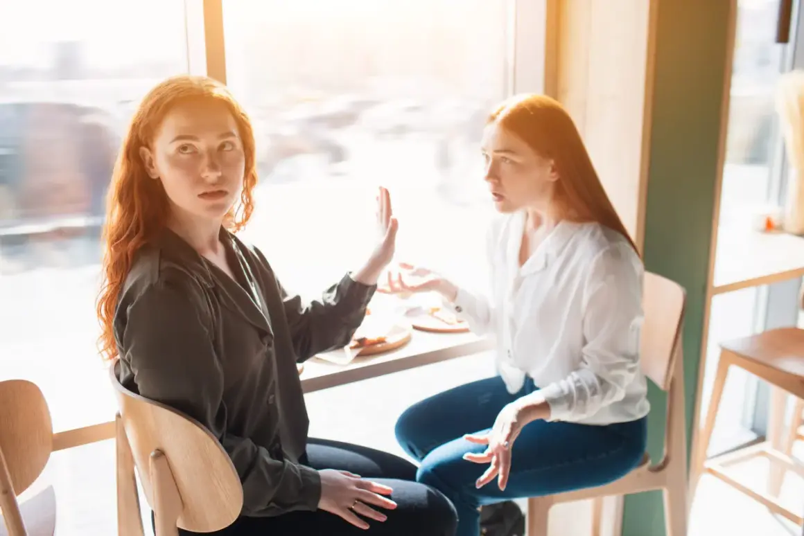 Zwei Frauen mit roten Haaren sitzen sich gegenüber. Eine hält die Hand hoch, als ob sie sagt: "Hör auf zu reden", wenn Kollegen nicht mit einem sprechen.