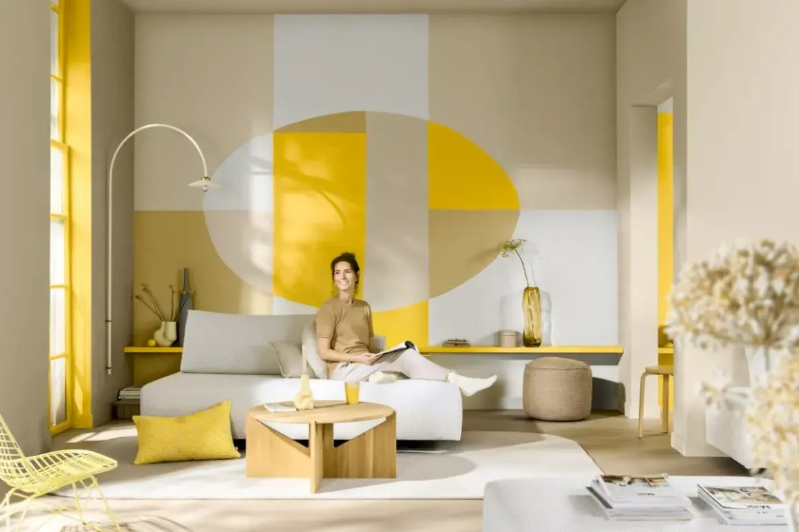 Eine Frau entspannt sich in einem hellen Wohnzimmer mit geometrischer Wandgestaltung. Welche Farbe passt zu Gelb? Hier: Beige, Grau und Weiß harmonieren perfekt.